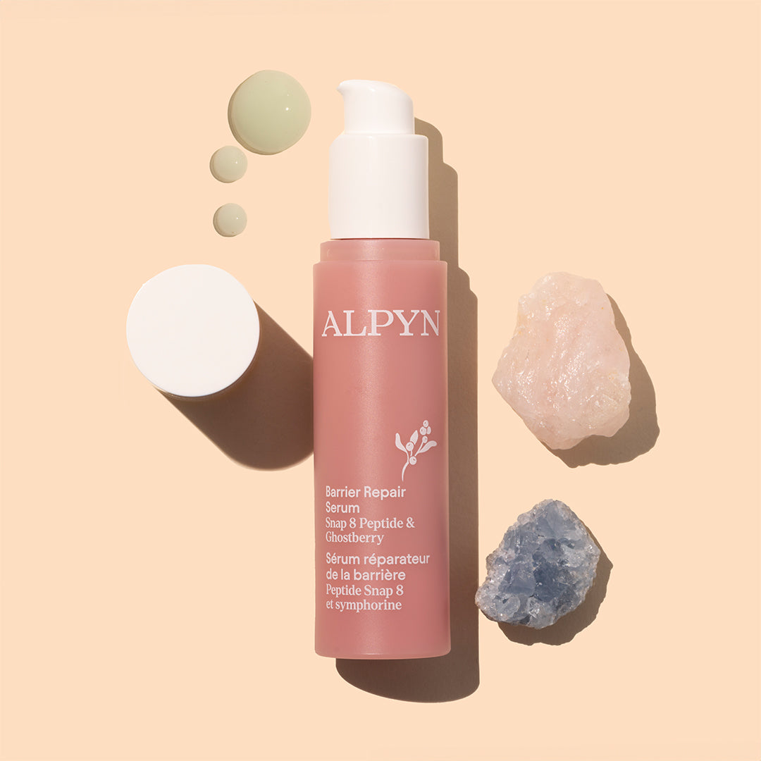Alpyn | Barrier Repair Serum | Styled Image with Swatch_Oct26 - dusty rose-pink pump bottle with pale green serum droplets, pink and blue crystals, and a white cap on a peach background