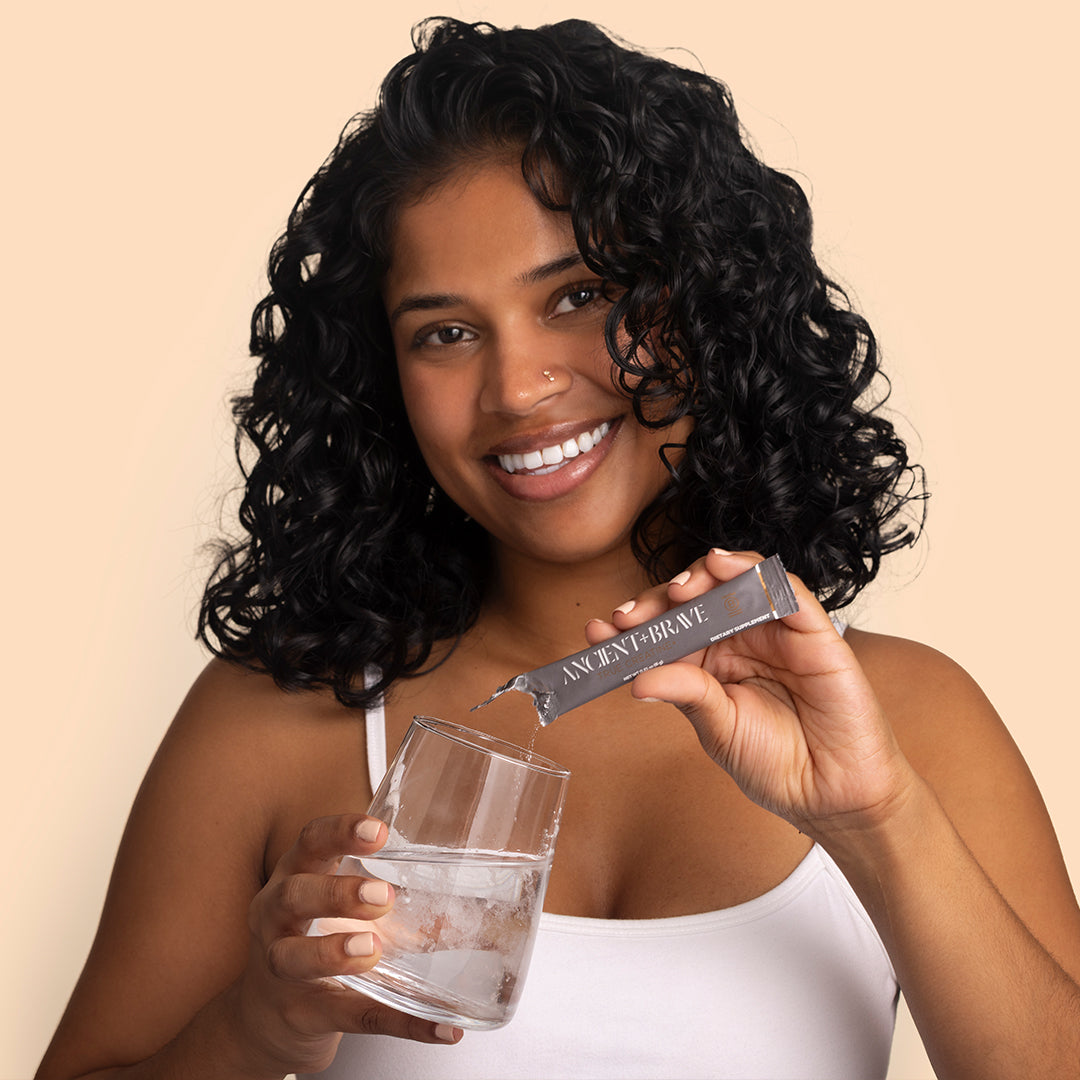 Ancient + Brave | True Creatine+ | Model Image - smiling person pouring powder into a glass of water against a beige background