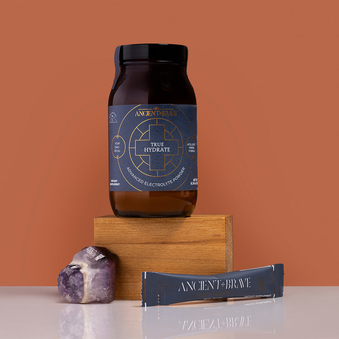 Ancient + Brave | True Hydrate | Product Image - jar displayed on wooden block with single-serve stick and crystal on a warm brown background