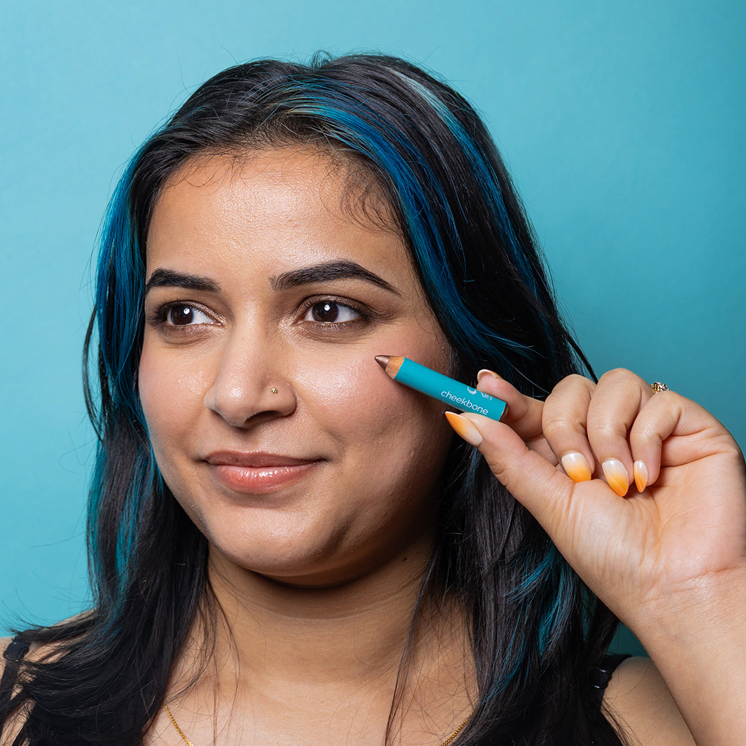 Cheekbone Beauty | Fearless Eyeshadow Mini Smokey Quartz Shade | Model Image - model with dark hair holding mini pencil in a shimmery bronze-brown shade against a turquoise blue background