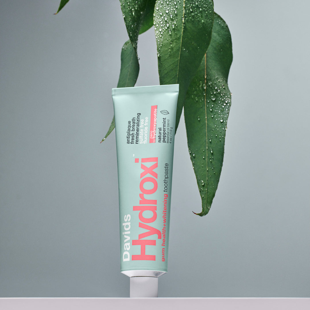 Davids | Hydroxi™ Gum Health + Whitening Nano Toothpaste - Peppermint | Lifestyle Image - mint green toothpaste tube with pink lettering displayed in front of dark green leaves covered in water droplets against a muted gray background