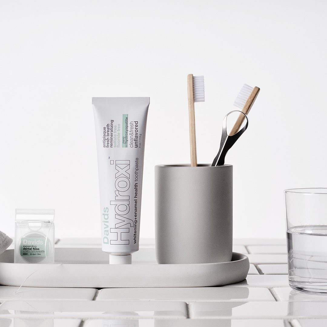 Davids | Hydroxi™ Whitening + Enamel Health Toothpaste Unflavored | Lifestyle Image - standing on a white bathroom tray beside dental floss, a toothbrush holder with toothbrushes and a tongue scraper, and a glass of water