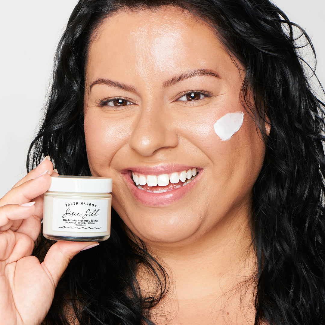 Earth Harbor | Siren Silk Bio-Retinol Hydration Cream | Model Image - smiling woman holding cream jar with a swatch of cream on her cheek