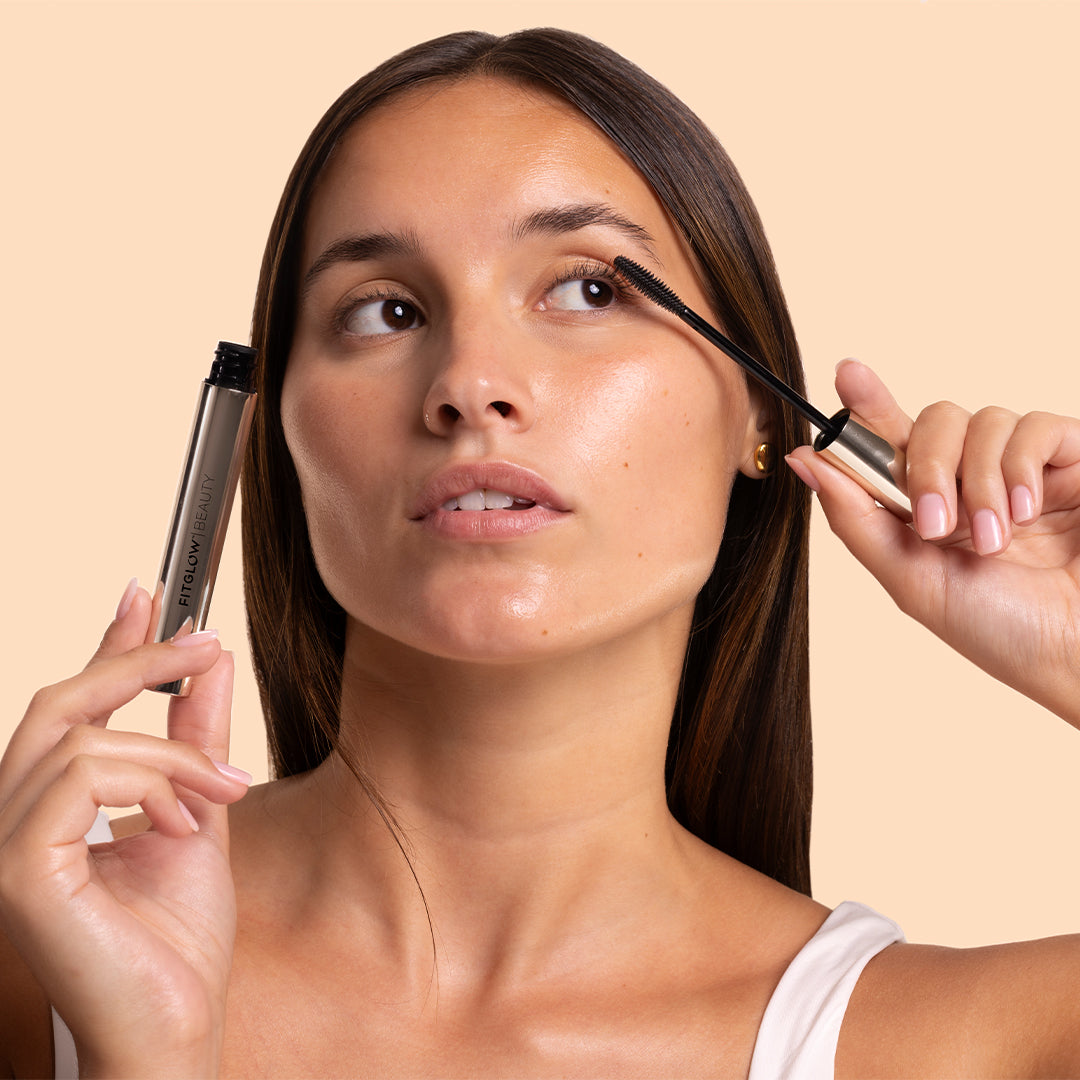 Fitglow Beauty | Lash Care Mascara | Model Image - model applying mascara while holding the silver mascara tube against a peach background
