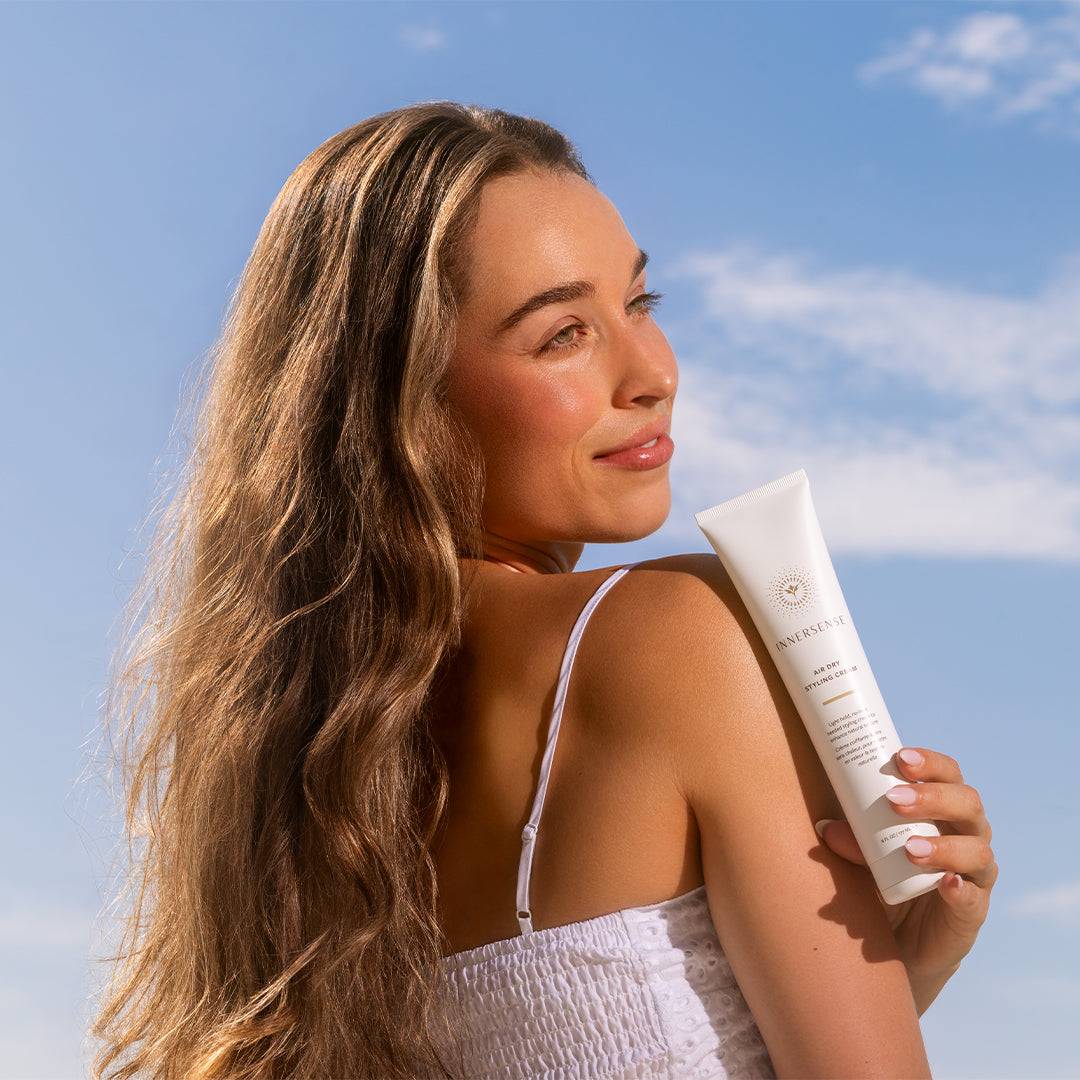 Innersense Organic Beauty | Air Dry Styling Cream | Model Image_Aug26 - person holding a white tube over their shoulder with long wavy hair against a bright blue sky background
