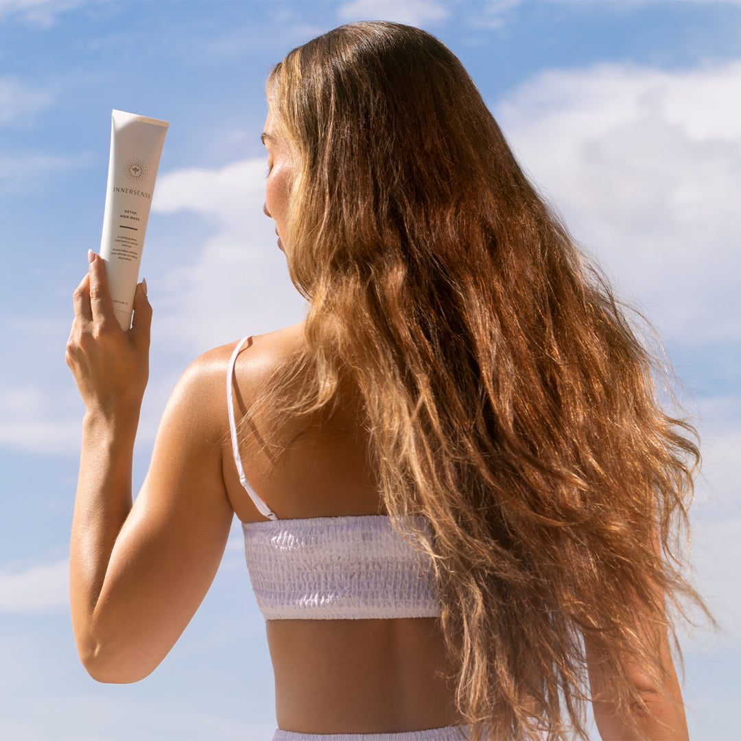 Innersense Organic Beauty | Detox Hair Mask | Model ImageA_Aug26 - back view of a person with long wavy hair holding a white tube of hair mask against a bright blue sky background