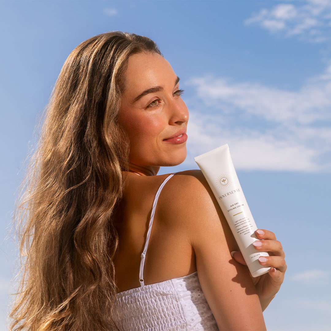 Innersense Organic Beauty | Overnight Air Dry Styling Cream | Model Image - Model holding cream bottle over her shoulder against a blue sky background