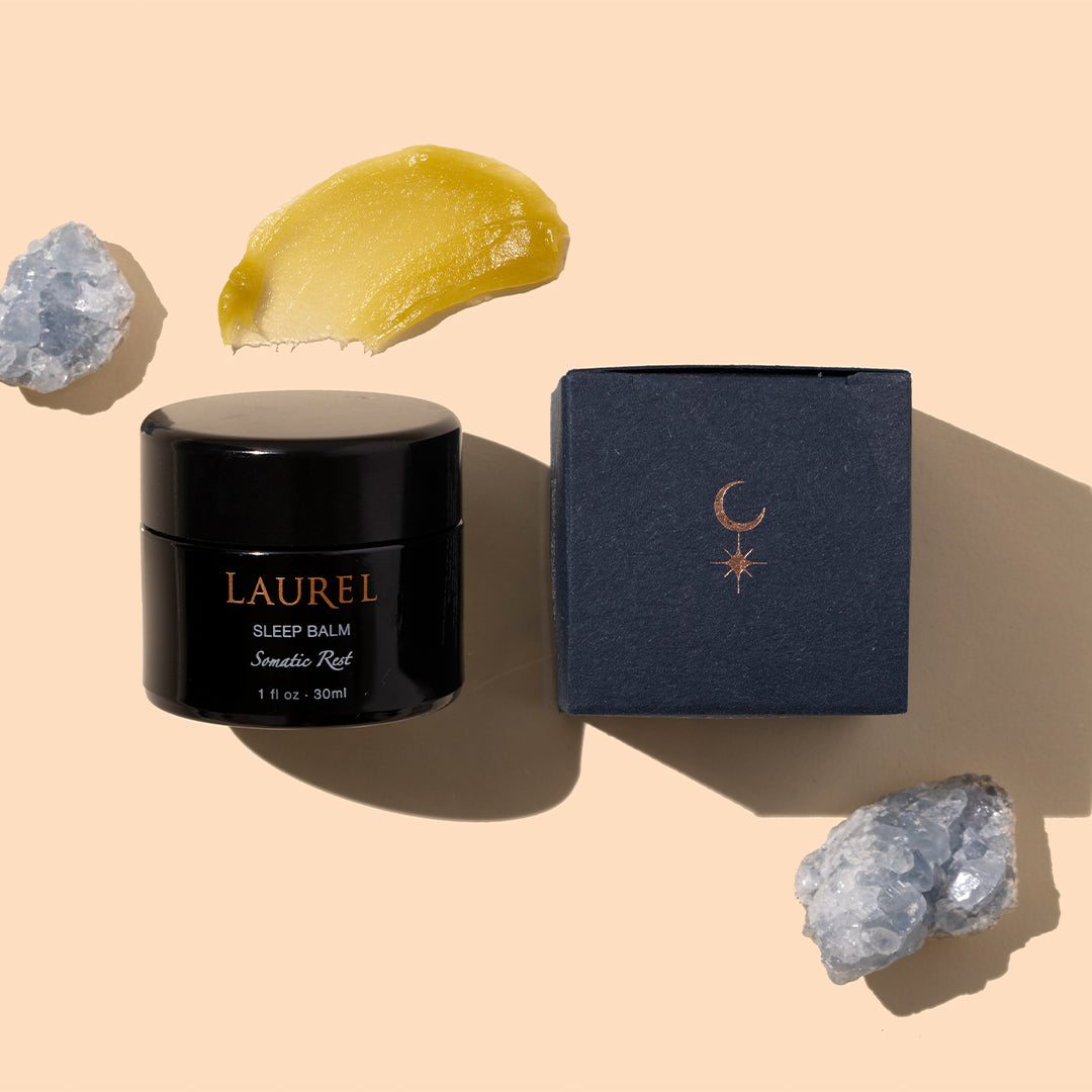 Laurel | Sleep Balm | Styled Image with Swatch_Sep26 - black jar with navy box and yellow balm swatch on a peach background with crystal accents