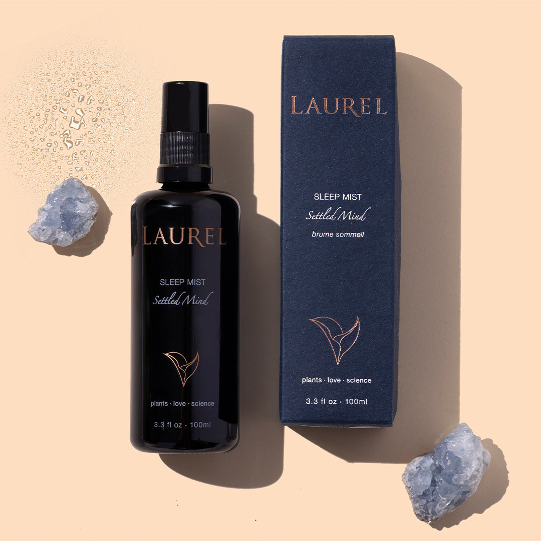 Laurel | Sleep Mist | Styled Image with Swatch_Sep26 - black spray bottle with navy box, water droplets, and crystal accents on a peach background