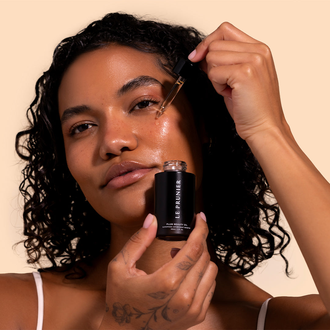 Le Prunier | Plum Beauty Oil | Model Image_Jul26 - model applying oil with a dropper while holding the matte black bottle beside the face