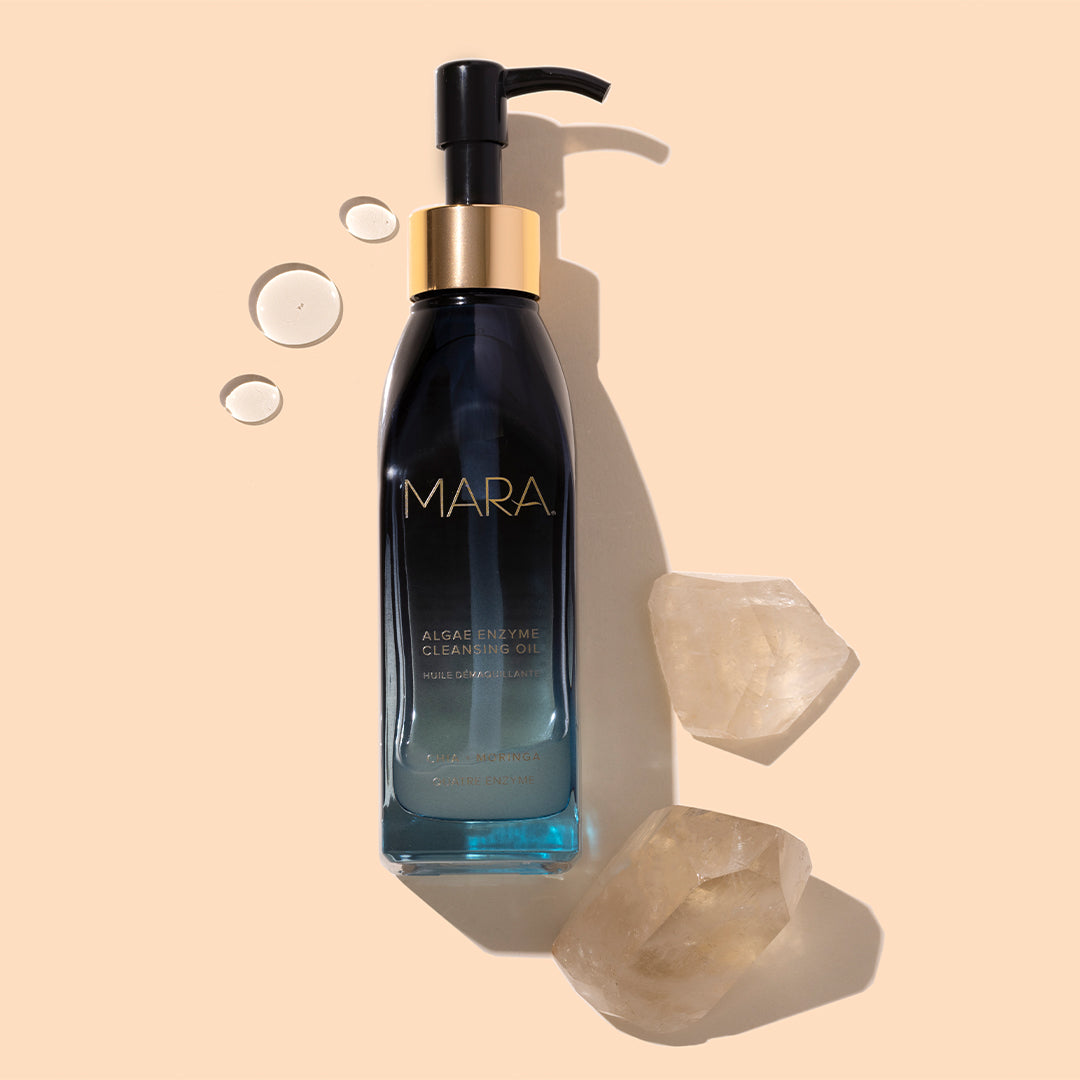 MARA | Algae Enzyme Cleansing Oil | Styled Image with Swatch_Jul26 - bottle with cleansing oil droplets and clear crystals on a beige background