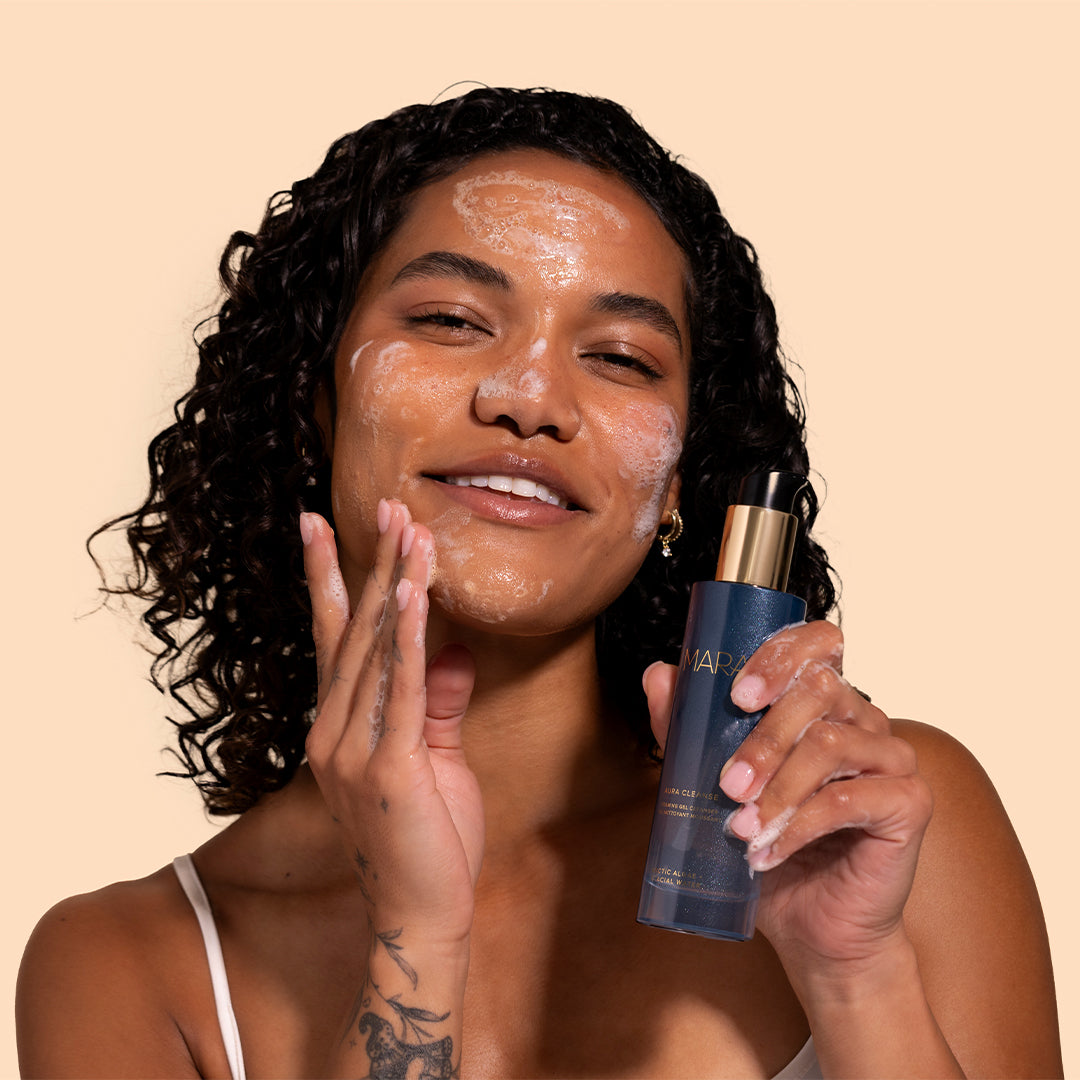 MARA | Aura Cleanse Foaming Gel Cleanser | Model Image_Jul26 - model cleansing while holding a blue bottle against a beige background