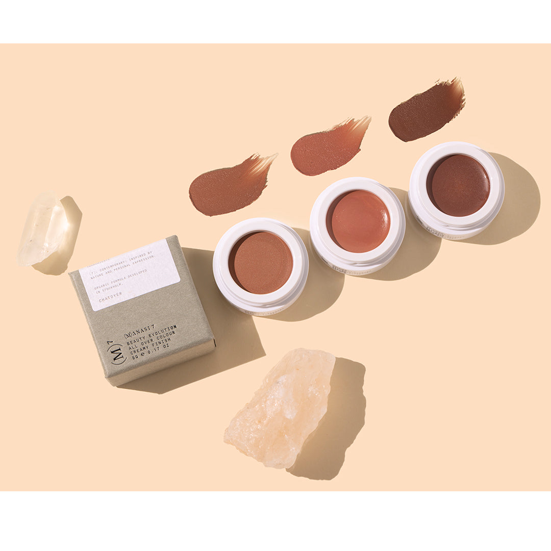 Manasi 7 | All Over Colour | Styled Image with Swatch_Oct26 - three creamy shades in warm caramel brown, terracotta rose, and deep chocolate brown with matching swatches, white pots, beige packaging, and pale crystals on a warm peach background