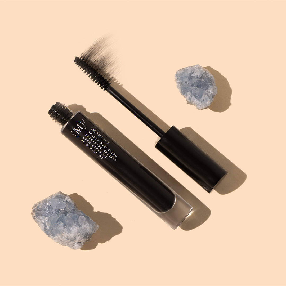 Manasi 7 | Precision Mascara | Styled Image with Swatch_Oct26 - black mascara tube and wand with icy blue crystals on a warm peach background