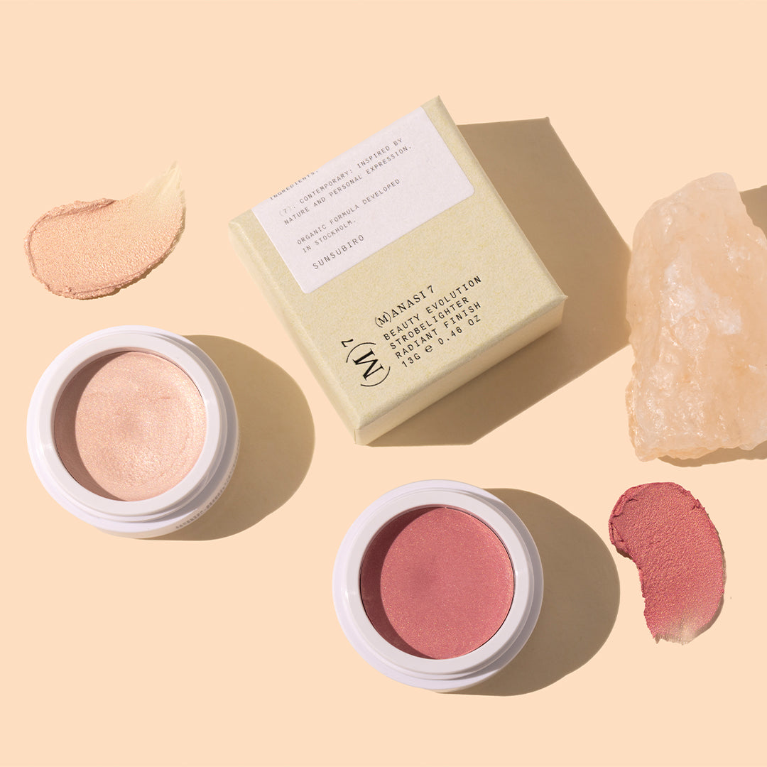 Manasi 7 | Strobelighter | Styled Image with Swatch_Oct26 - champagne-peach and rosy pink highlighters with matching shimmery swatches, white packaging, a pale beige box, and a blush-pink crystal on a warm peach background