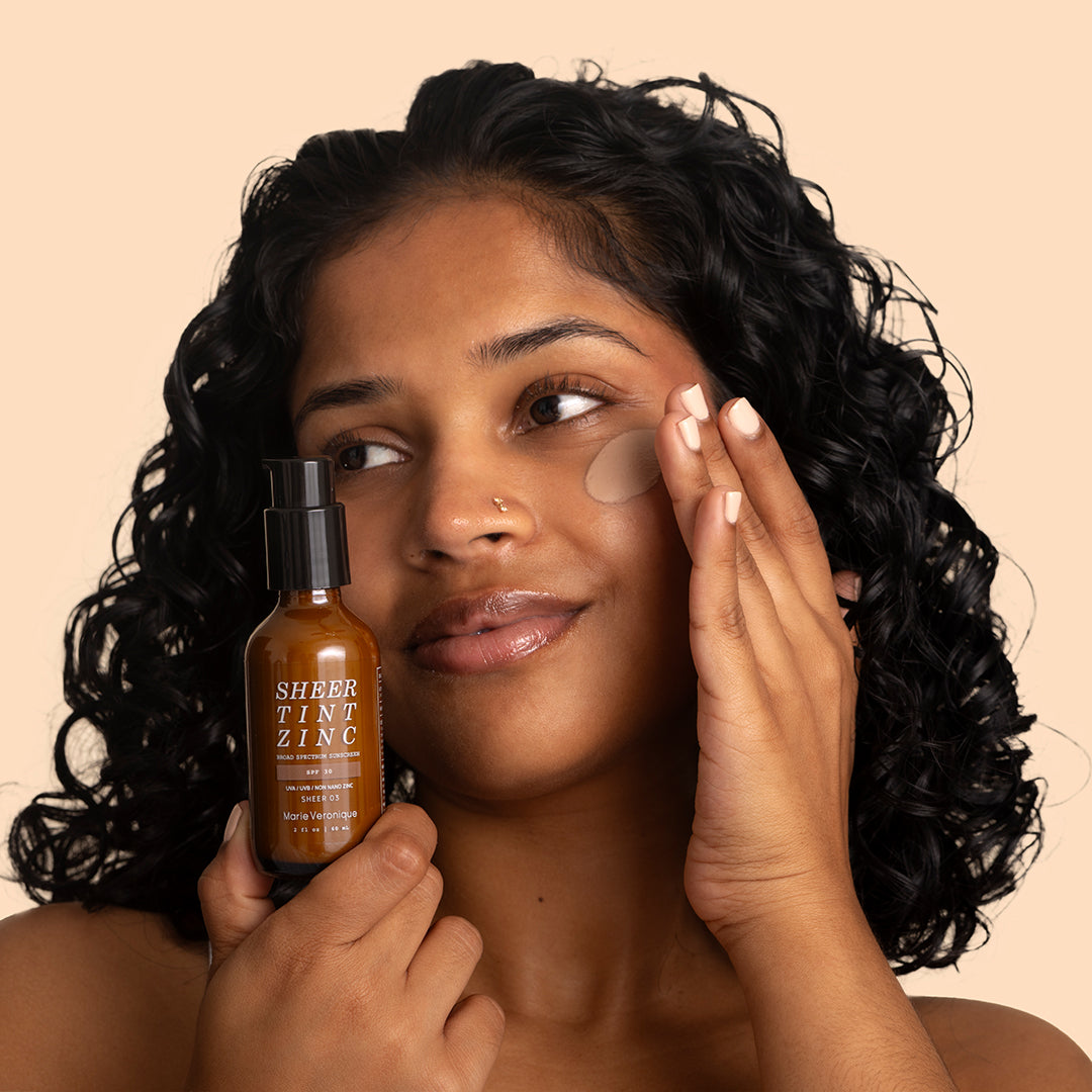 Marie Veronique | Sheer Tint Zinc SPF 30 | Model Image - person applying tint to cheek while holding the bottle against a beige background