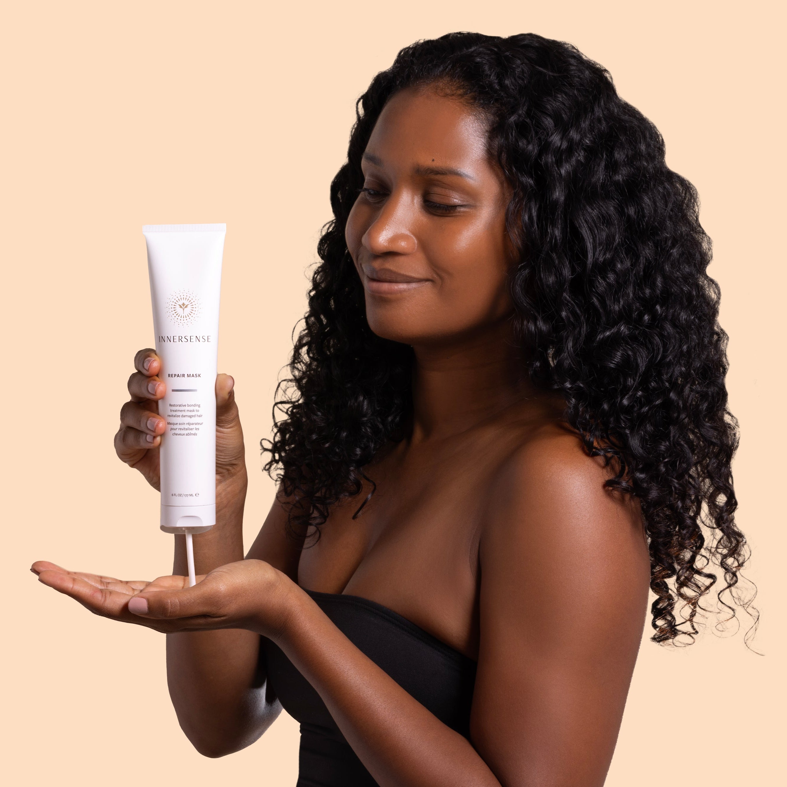 May Lindstrom Skin | The Problem Solver | Model ImageA_Aug26 - model with curly black hair dispensing repair mask from a white tube into her palm against a light beige background