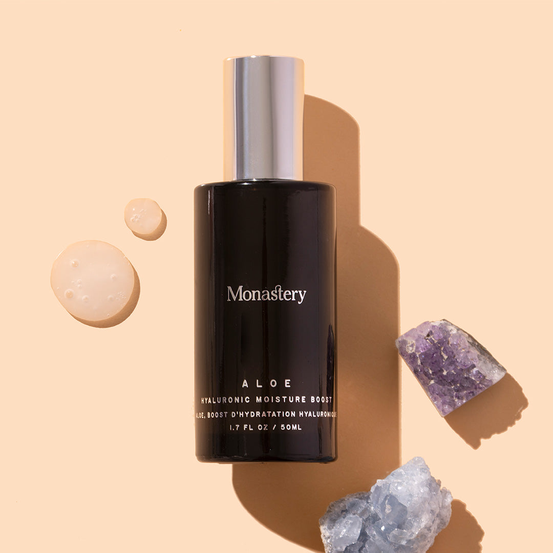 Monastery | Aloe Hyaluronic Moisture Boost | Styled image with swatch, dark bottle with silver cap on peach background with white gel swatch and two crystals