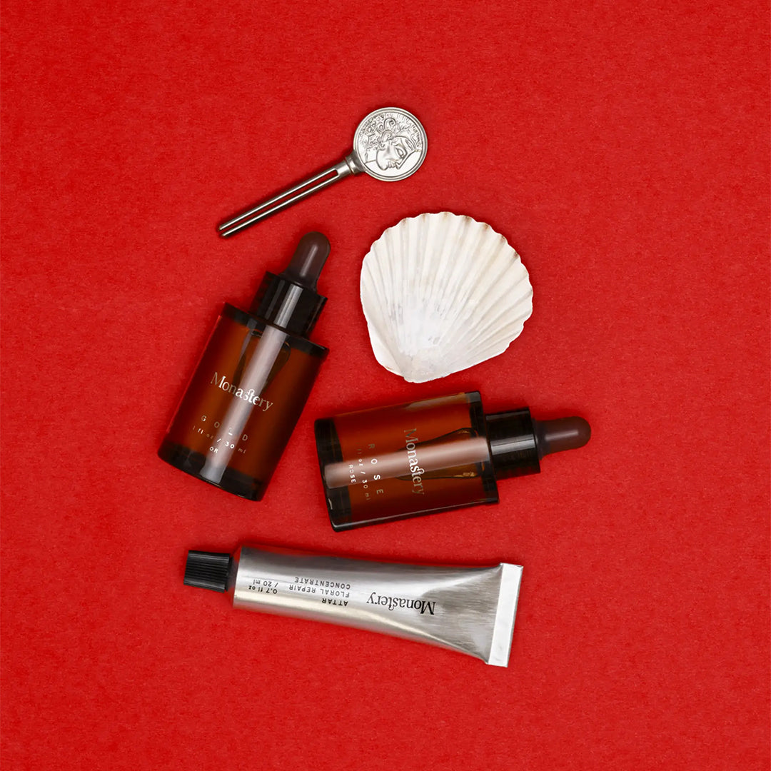 Monastery | Essentials Kit | Styled Image - skincare set with Rose and Gold oils, Attar Floral Repair Concentrate, and Monastery coin logo key on a red background