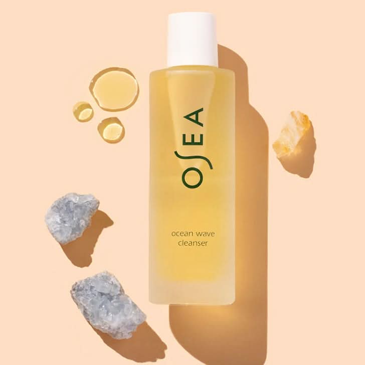 OSEA | Ocean Wave Cleanser | Styled image flat lay with swatch, A bottle of OSEA Ocean Wave Cleanser with a white cap, filled with pale yellow liquid, set against a warm peach background with decorative crystals and liquid droplets.