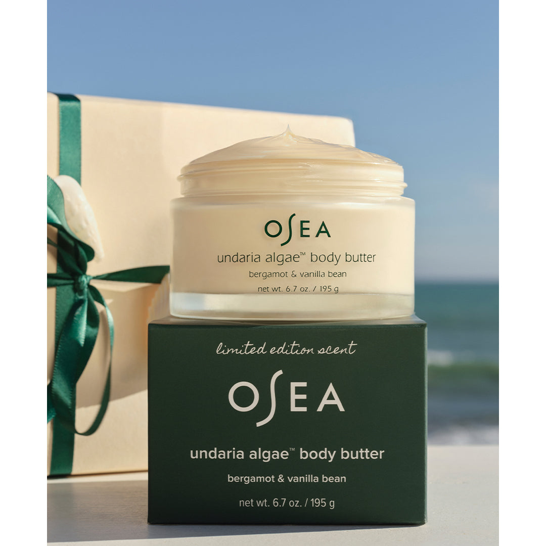 OSEA | Undaria Algae Body Butter | Packaging Image - open body butter jar displayed on its green box beside a ribbon-wrapped gift with an ocean backdrop.