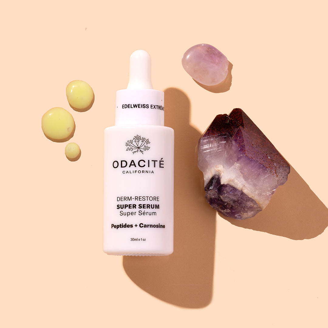 Odacité | Edelweiss Extrême Derm-Restore Super Serum | Product Image with Swatch - bottle with dropper on a warm beige background, surrounded by yellow serum droplets and stones