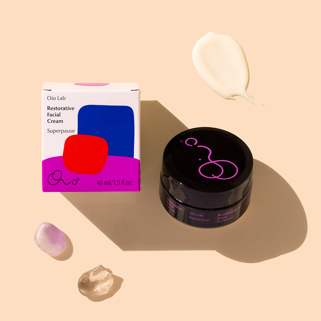Oio Lab | Superpause Restorative Facial Cream | Product Image - woman holding black cream jar with box, oil drops, and crystals against a beige background