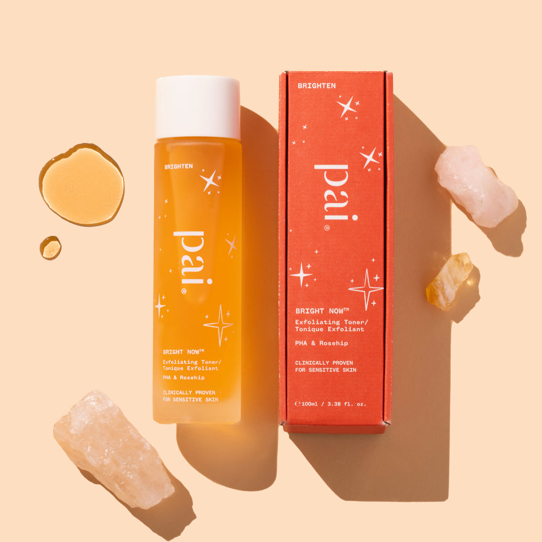 Pai Skincare | Bright Now PHA & Rosehip Exfoliating Toner | Product Image with Swatch - bottle with box, liquid drops, and crystals on a warm neutral background
