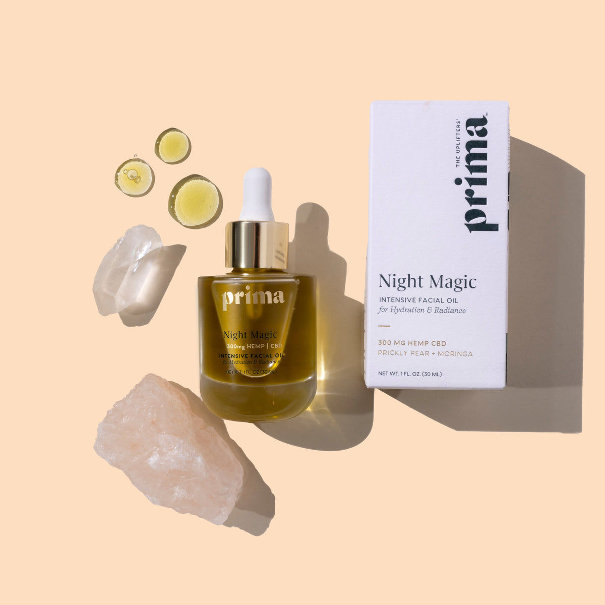 Prima | Night Magic Intensive Facial Oil | Styled image flat lay with swatch | Clear bottle with a white dropper cap next to its white product box, styled with crystal stones and small oil droplets on a warm beige background.