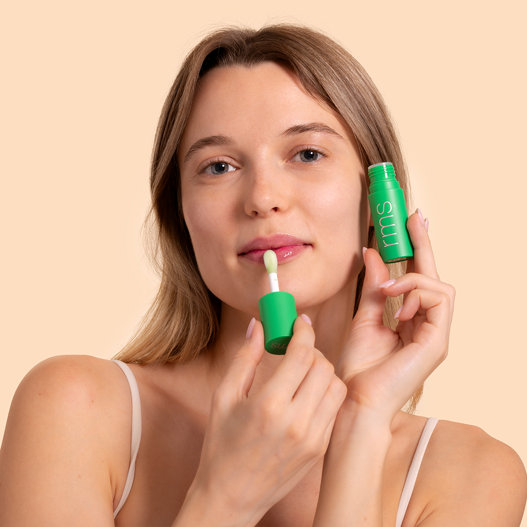 RMS Beauty | Chameleon Lip Oil | Model Image - woman applying lip oil from a tube against a beige background