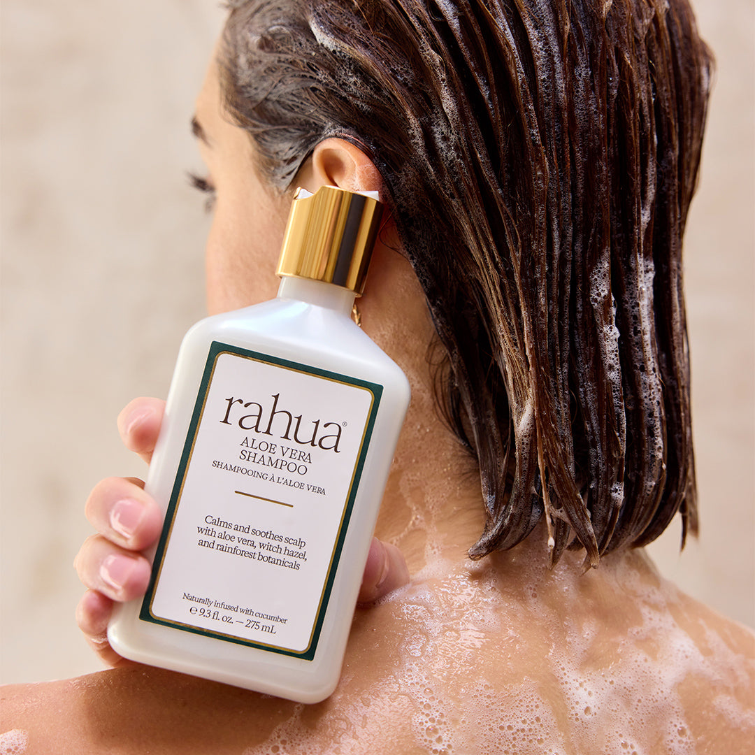 Rahua | Aloe Vera Shampoo | Model Image - model holding a white shampoo bottle with a gold cap while washing hair