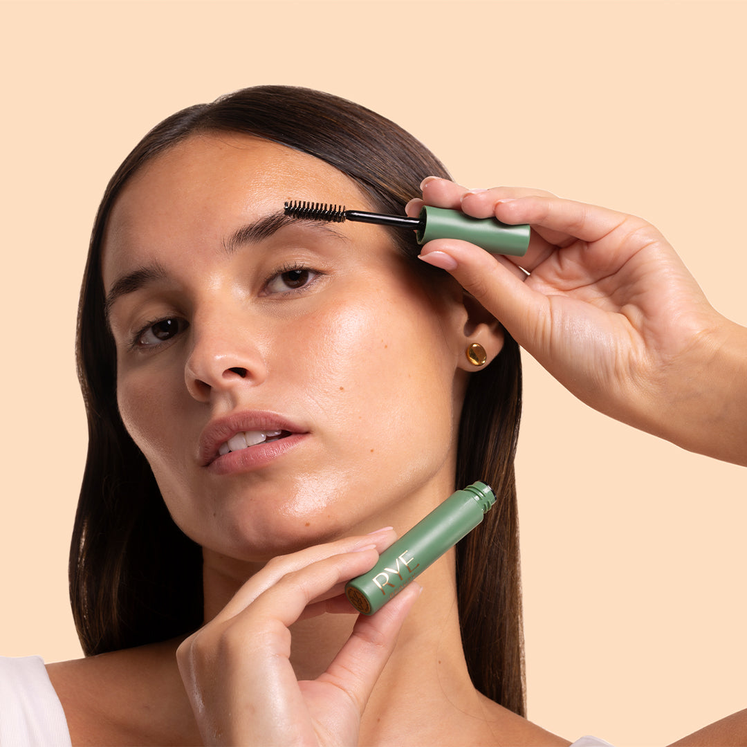 Rye Beauty | Brow Grooming Serum | Model Image_Oct26 - model applying brow serum from a sage green tube against a peach background