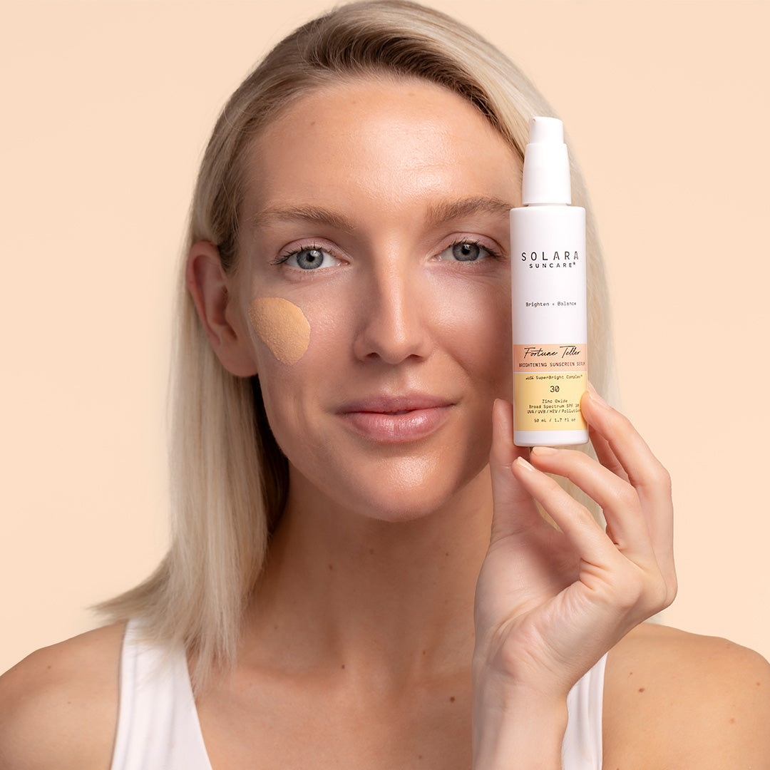 Solara Fortune Teller with model with swatch, Model on peach background with product swatch on skin, holding white product bottle near face