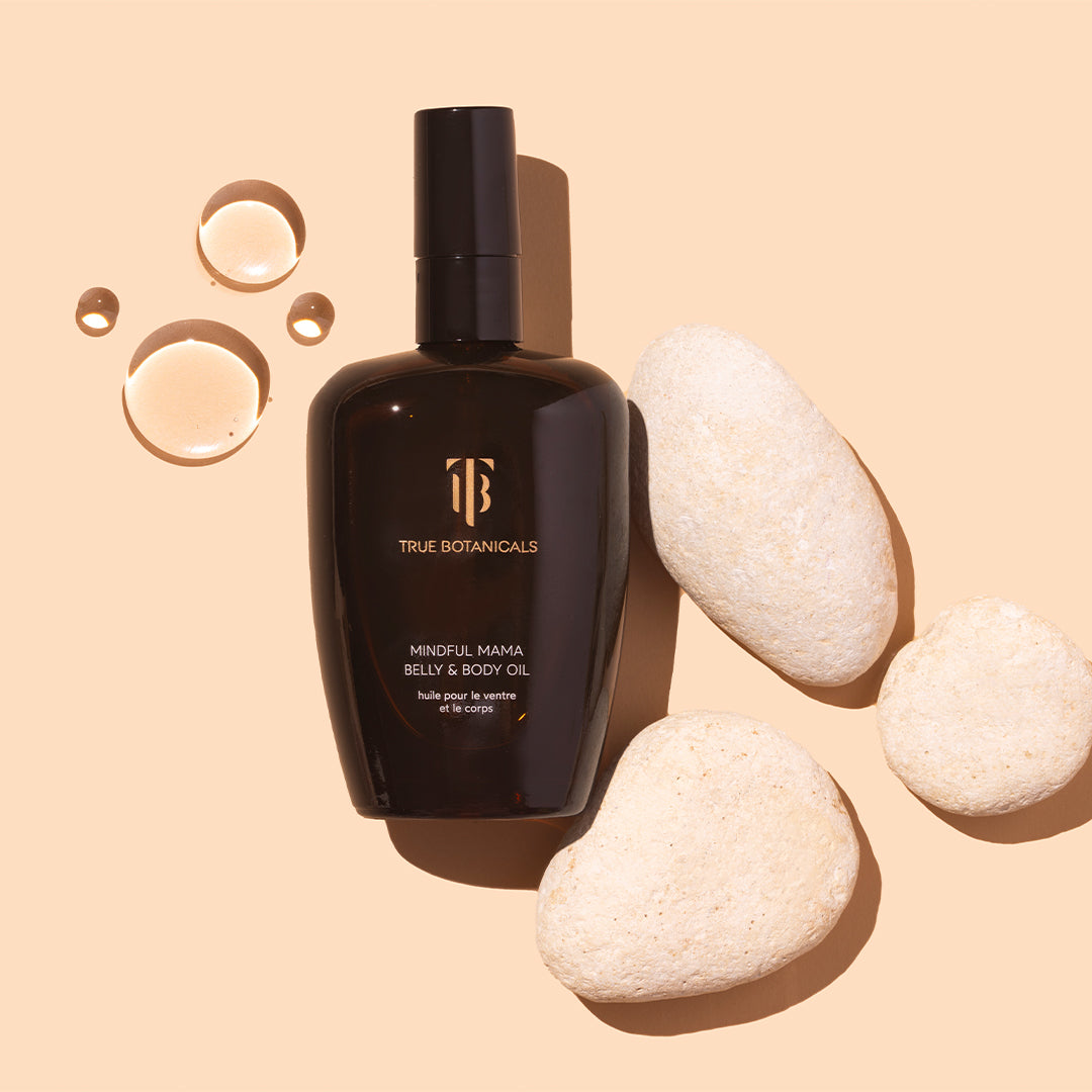 True Botanicals | Mindful Mama Belly & Body Oil | Styled Image with Swatch - dark amber bottle with oil droplets and smooth stones on a beige background