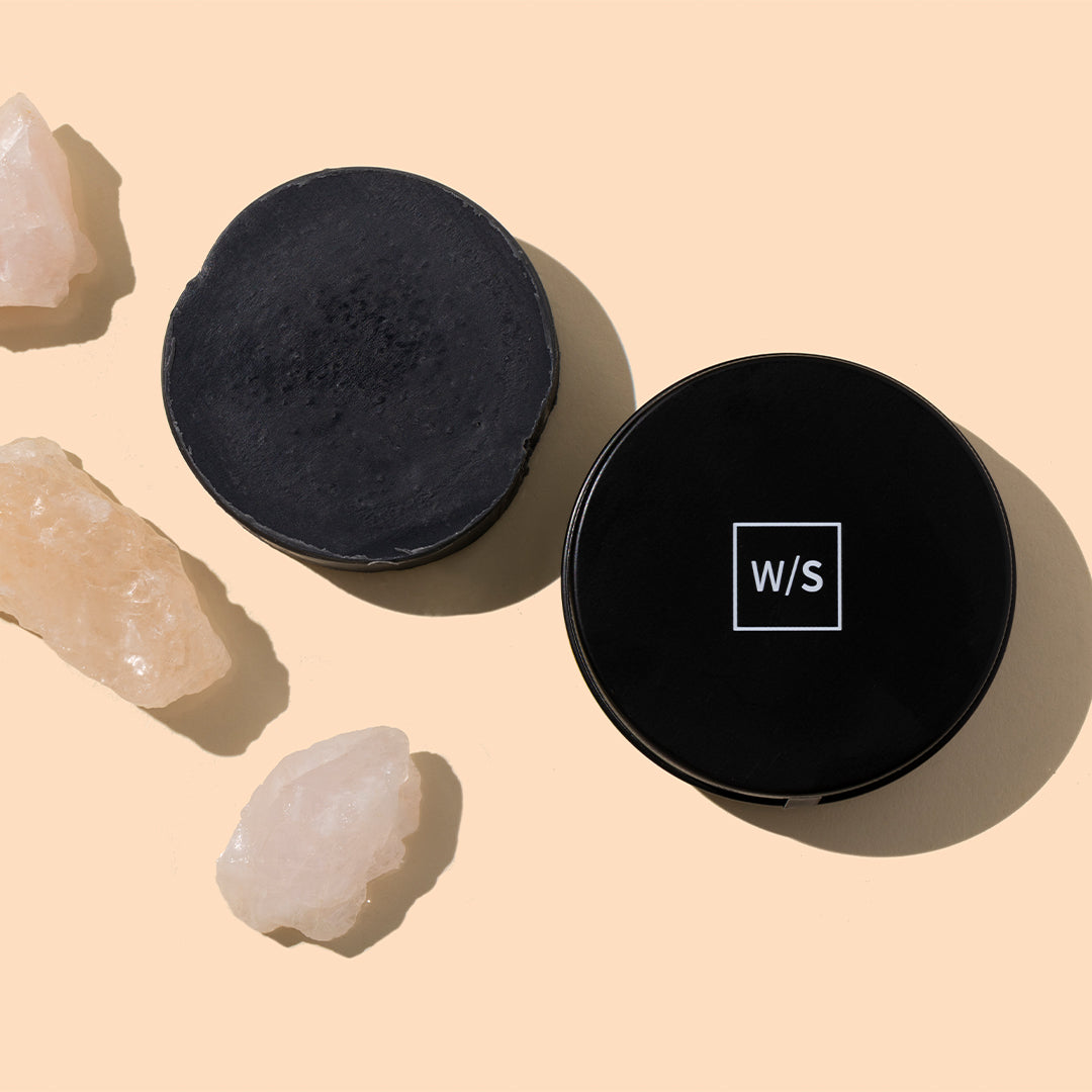withSimplicity | Charcoal Cleanser | Styled Image_Sep26 - charcoal cleanser in a black round container with lid beside the product, styled with white crystals on a peach background
