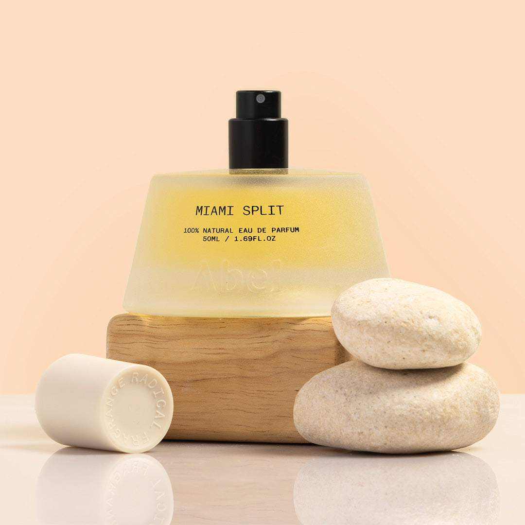 Abel | Miami Split Eau de Parfum | Styled Image | Frosted glass erfume bottle with black cap sitting on wooden black with two rocks, styled photograph