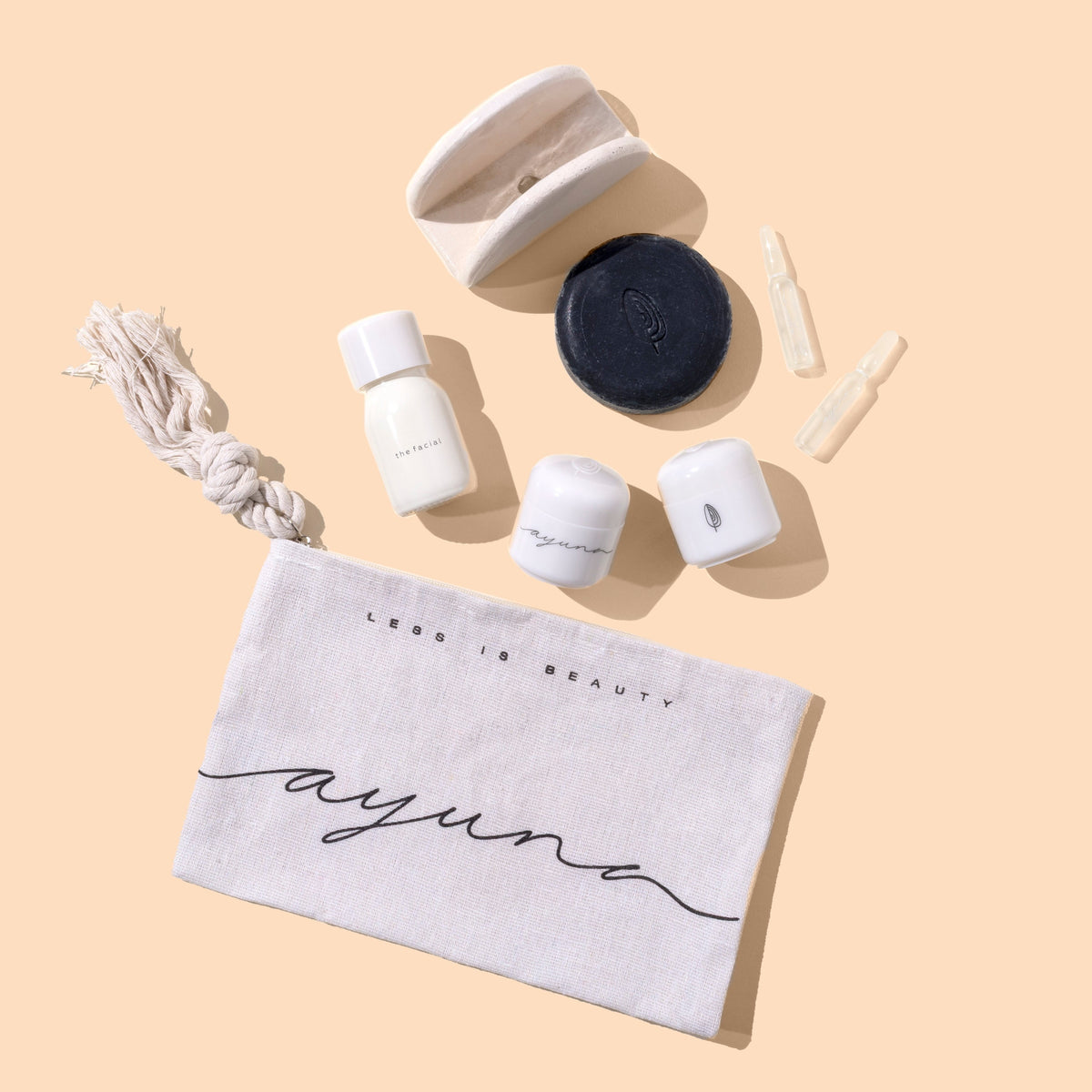 Ayune skincare set laid out on a warm beige background, including a white cotton pouch with black script logo, a white cord, several small white cosmetic containers, a round black lid, and two clear applicator tips. The pouch reads 'HERE IS BEAUTY ayune'.