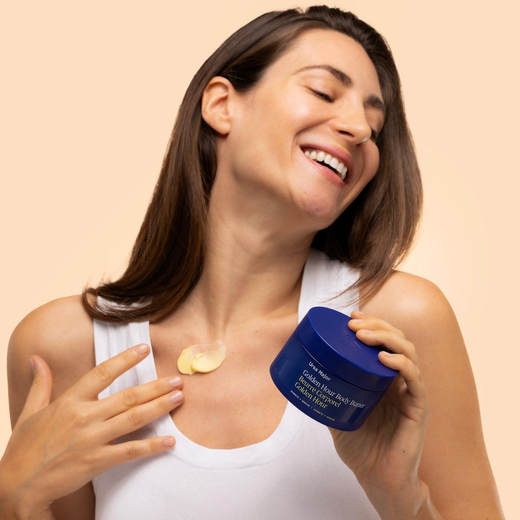 Ursa Major Golden Hour Body Butter - Woman applying a product from a blue jar on a beige background