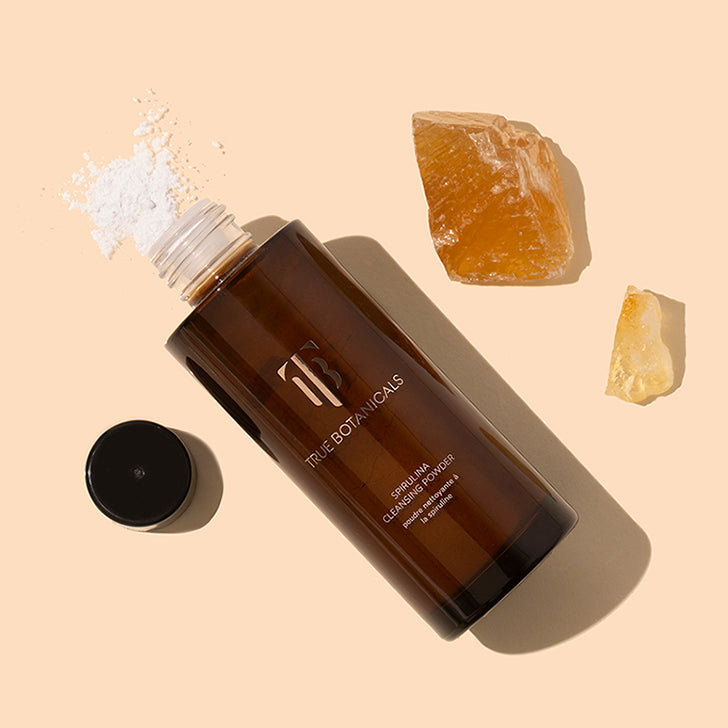 True Botanicals | Spirulina Cleansing Powder | Styled image with swatch | Brown bottle with dark cap open on peach background with white powder sprinkling out