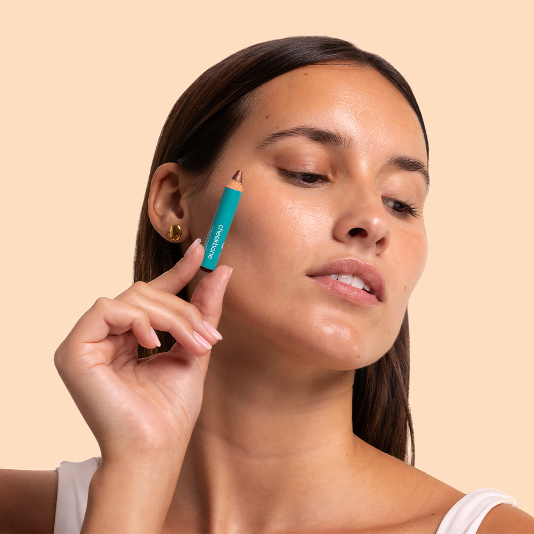 Cheekbone Beauty | Fearless Eyeshadow Mini | Model Image - model holding mini pencil near her eye against a peach background