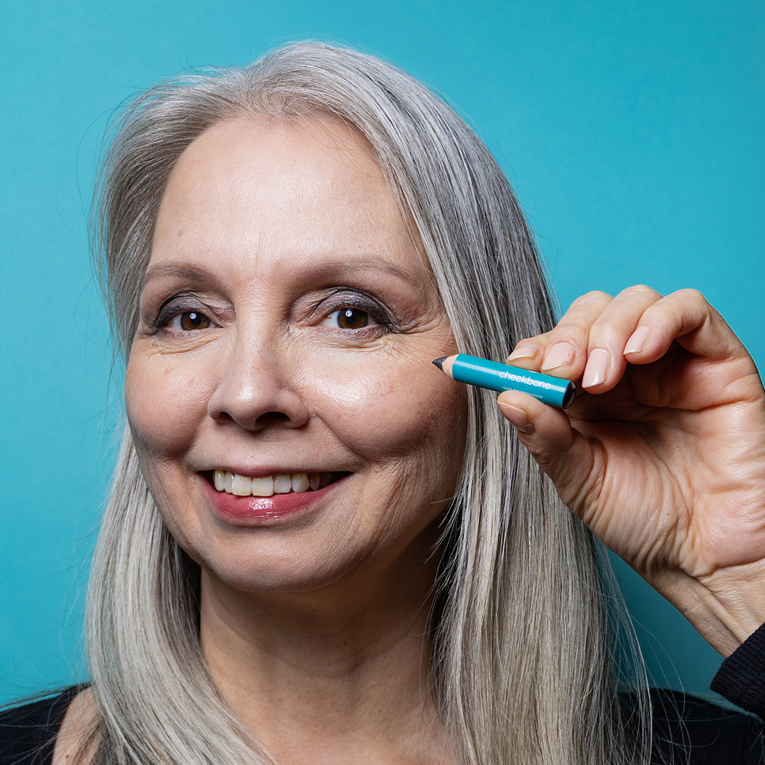 Cheekbone Beauty | Fearless Eyeshadow Mini Onyx Shade | Model Image - model with silver-gray hair holding mini pencil in a smoky charcoal shade against a turquoise blue background