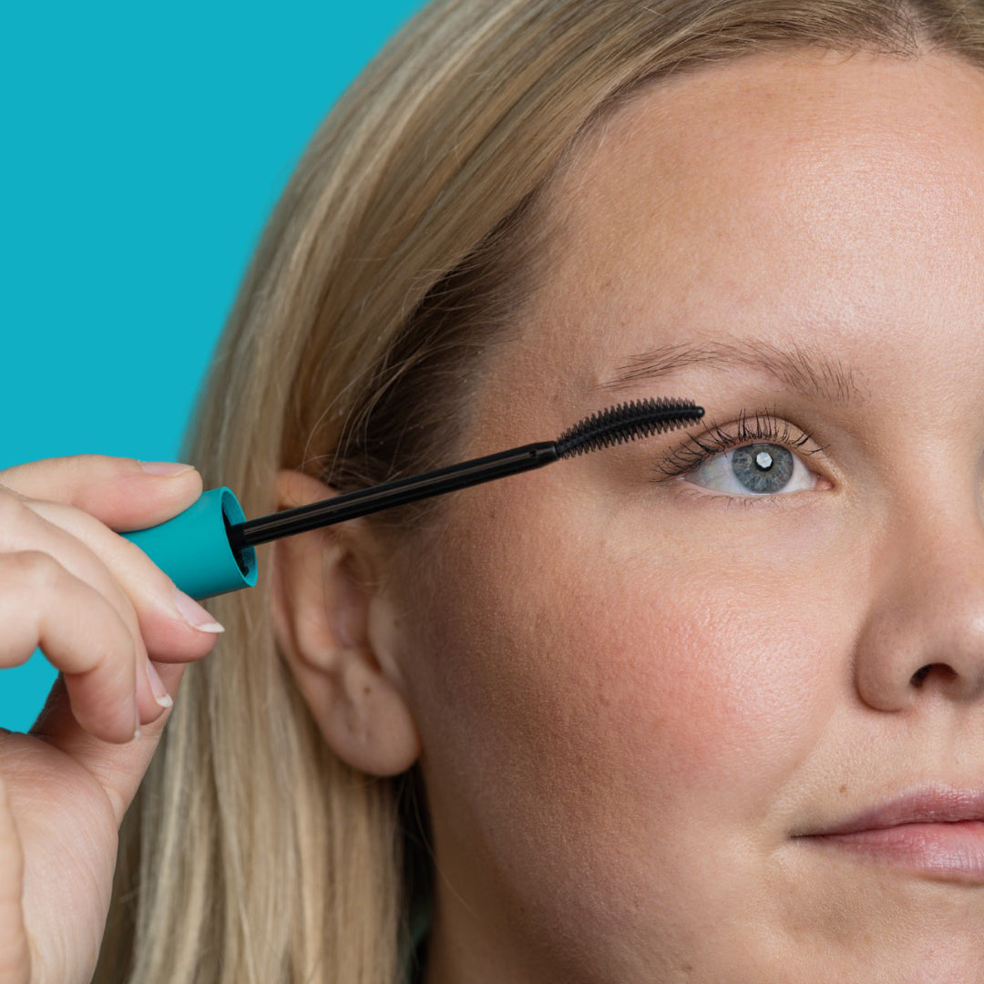 Cheekbone Beauty | Uprise Mascara | Model Image - model applying mascara to lashes on a turquoise blue background

