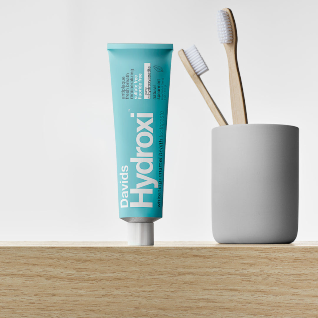 Davids | Hydroxi™ Whitening + Enamel Health Toothpaste - Spearmint | Product Image - blue toothpaste tube  beside bamboo toothbrushes in a bathroom setting