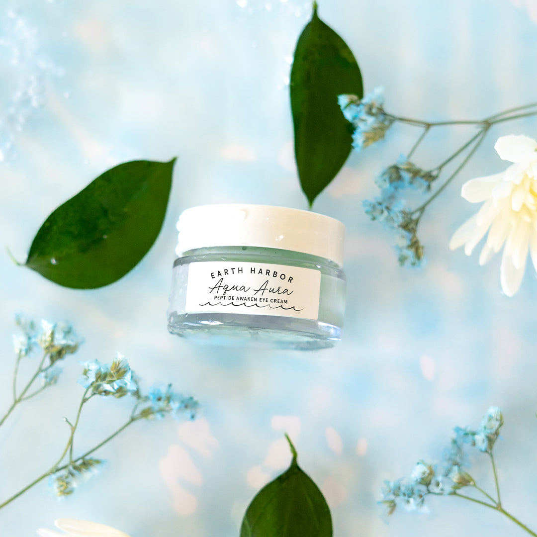 Earth Harbor | Aqua Aura Peptide Awaken Eye Cream | Lifestyle Image - glass jar surrounded by green leaves and delicate flowers on a light blue background
