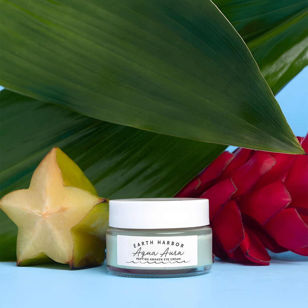 Earth Harbor | Aqua Aura Peptide Awaken Eye Cream | Lifestyle Image 2 - glass jar displayed with starfruit, tropical flowers, and green leaves