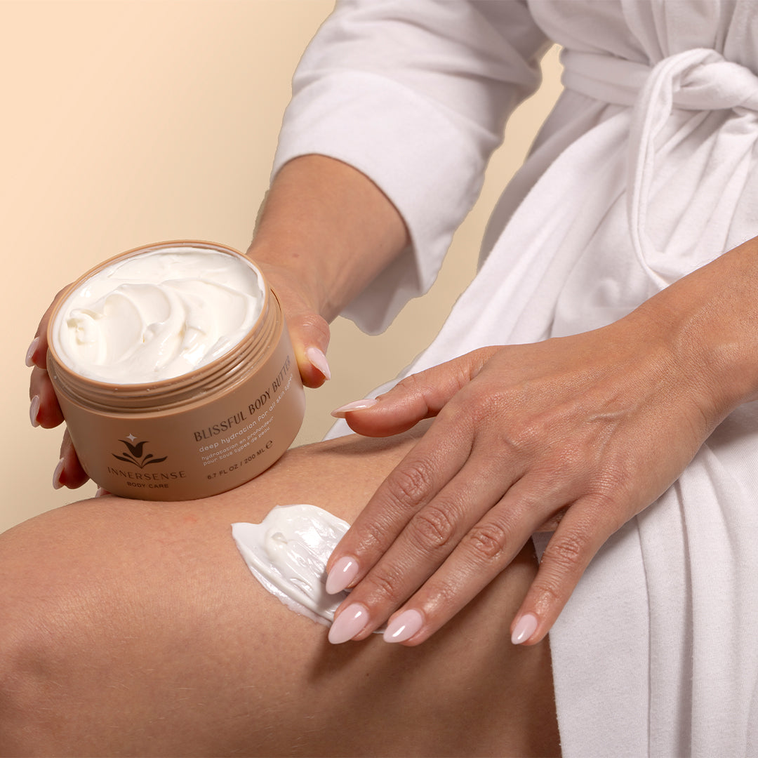 Innersense Organic Beauty | Blissful Body Butter | Model Image_Aug26 - close-up of hands applying body butter from an open jar onto a leg while wearing a white robe against a light beige background