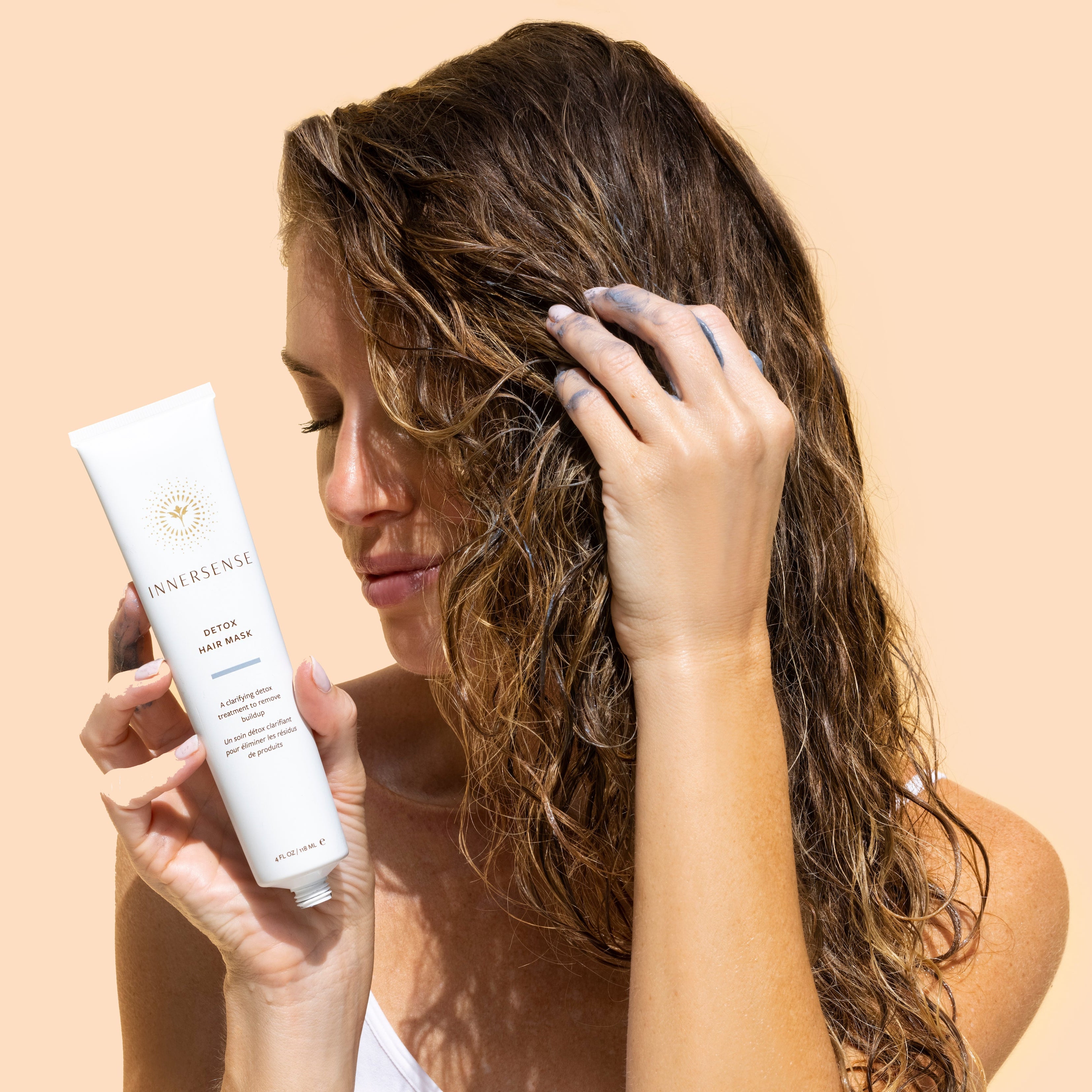 Innersense Organic Beauty |
Detox Hair Mask| Model Image_Aug26 - person with wet hair holding an open tube of mask while applying the dark gray mask to their hair against a soft beige background