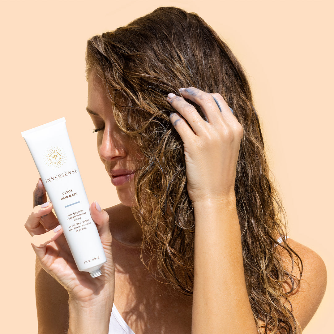 Innersense Organic Beauty | Overnight Detox Hair Mask | Model Image - model tube beside damp hair while applying the gray mask to her scalp on a beige background