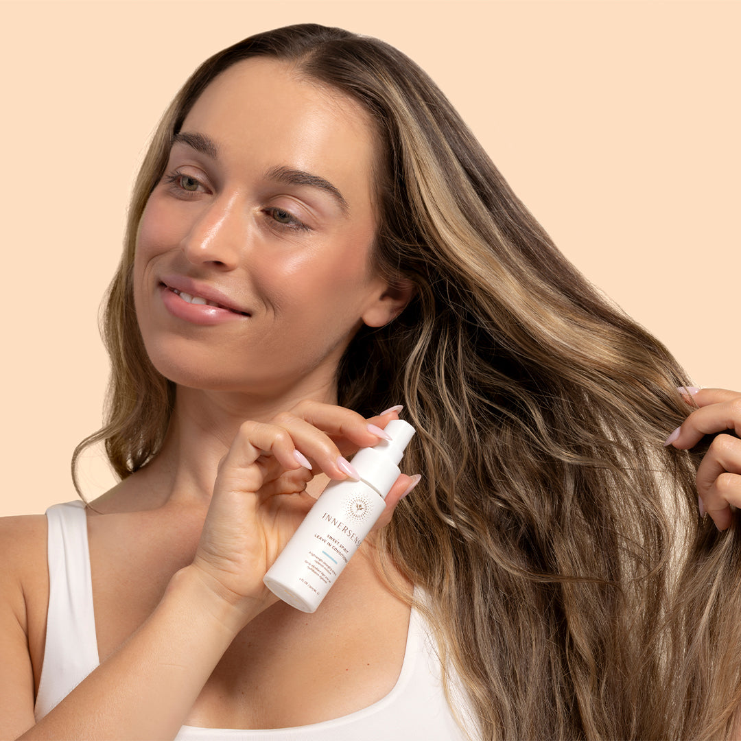 Innersense Organic Beauty | Sweet Spirit Leave-In Conditioner | Model Image_Aug26 - smiling model holding a white bottle while lifting a section of her long hair against a light beige background