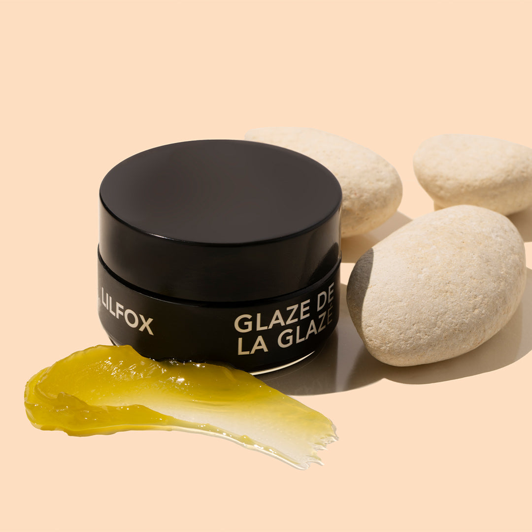 LILFOX | Glaze | Styled Image with Swatch_Oct26 - black jar with a golden-yellow product swatch and cream-colored stones on a peach background